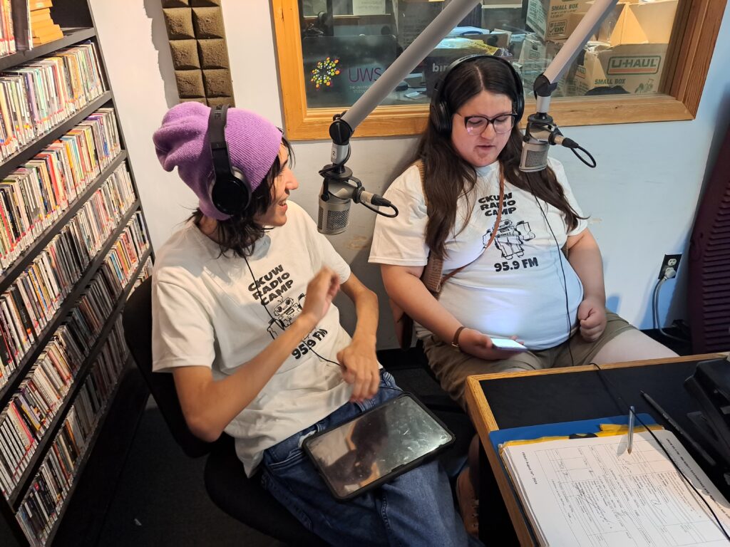 Two youth are sitting in a radio studio talking into microphones. They are wearing t-shirts that say 'Radio Camp'.